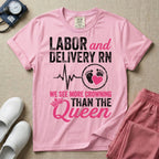 A funny graphic t-shirt for a Labor and Delivery unit featuring distressed white and pink typography that reads 'LABOR and DELIVERY RN' above a heartbeat line connecting to baby footprints and a pink heart, followed by the humorous phrase 'WE SEE MORE CROWNING THAN THE Queen' with a pink crown illustration.
