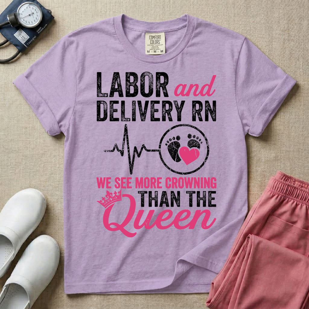 A funny graphic t-shirt for a Labor and Delivery unit featuring distressed white and pink typography that reads 'LABOR and DELIVERY RN' above a heartbeat line connecting to baby footprints and a pink heart, followed by the humorous phrase 'WE SEE MORE CROWNING THAN THE Queen' with a pink crown illustration.