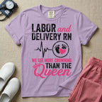 A funny graphic t-shirt for a Labor and Delivery unit featuring distressed white and pink typography that reads 'LABOR and DELIVERY RN' above a heartbeat line connecting to baby footprints and a pink heart, followed by the humorous phrase 'WE SEE MORE CROWNING THAN THE Queen' with a pink crown illustration.