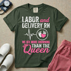 A funny graphic t-shirt for a Labor and Delivery unit featuring distressed white and pink typography that reads 'LABOR and DELIVERY RN' above a heartbeat line connecting to baby footprints and a pink heart, followed by the humorous phrase 'WE SEE MORE CROWNING THAN THE Queen' with a pink crown illustration.