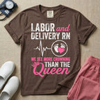 A funny graphic t-shirt for a Labor and Delivery unit featuring distressed white and pink typography that reads 'LABOR and DELIVERY RN' above a heartbeat line connecting to baby footprints and a pink heart, followed by the humorous phrase 'WE SEE MORE CROWNING THAN THE Queen' with a pink crown illustration.
