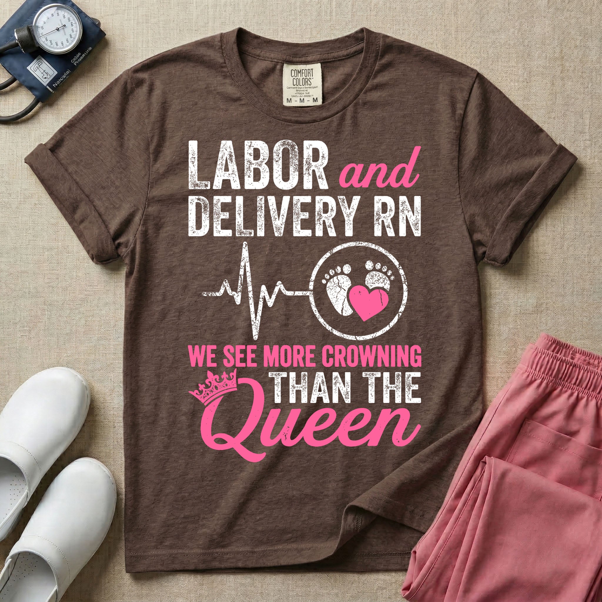 A funny graphic t-shirt for a Labor and Delivery unit featuring distressed white and pink typography that reads 'LABOR and DELIVERY RN' above a heartbeat line connecting to baby footprints and a pink heart, followed by the humorous phrase 'WE SEE MORE CROWNING THAN THE Queen' with a pink crown illustration.