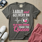 A funny graphic t-shirt for a Labor and Delivery unit featuring distressed white and pink typography that reads 'LABOR and DELIVERY RN' above a heartbeat line connecting to baby footprints and a pink heart, followed by the humorous phrase 'WE SEE MORE CROWNING THAN THE Queen' with a pink crown illustration.