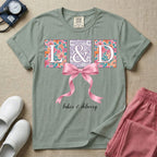 A graphic t-shirt for a Labor and Delivery unit featuring the white serif letters 'L&D' set inside colorful floral square panels, accented with a large graphic pink bow across the middle and the text 'labor & delivery' in delicate white script at the bottom.