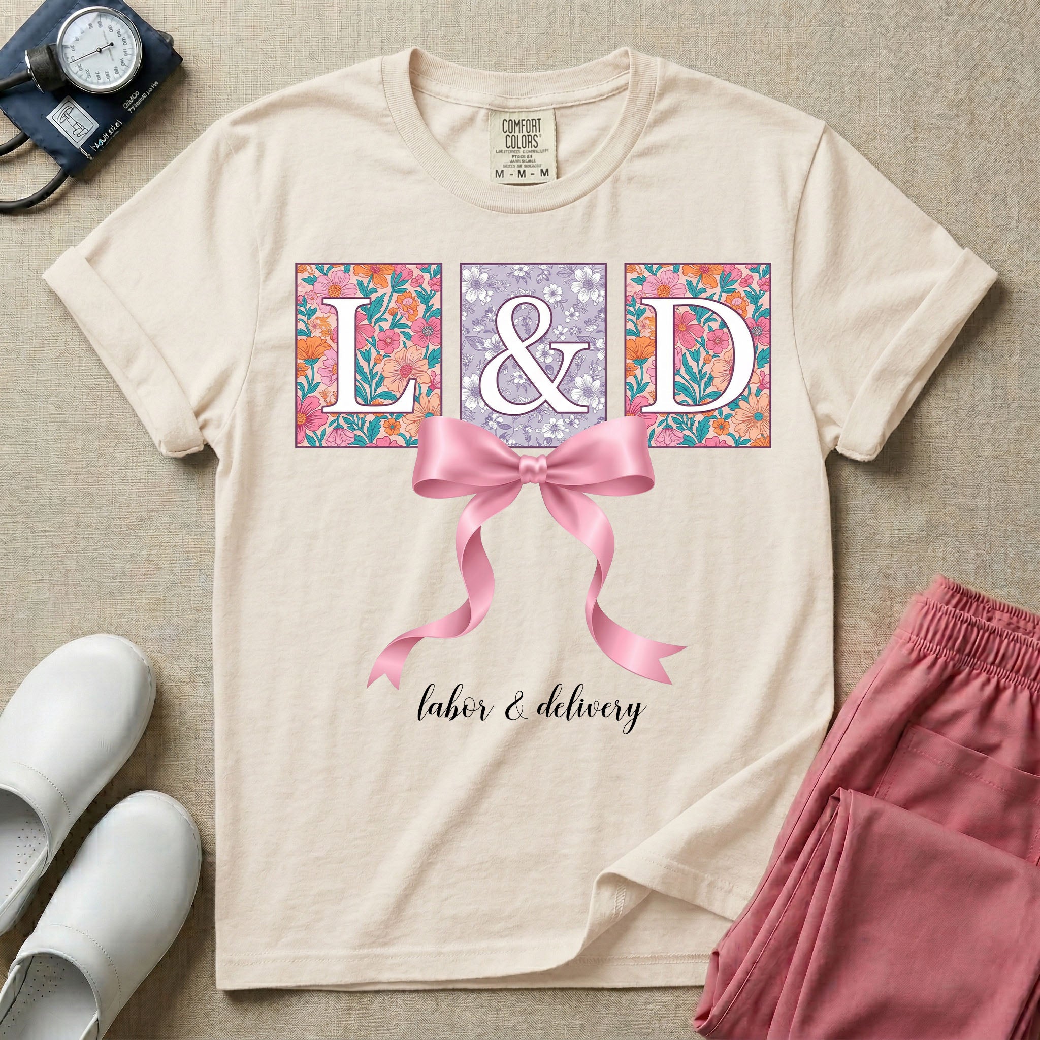 A graphic t-shirt for a Labor and Delivery unit featuring the white serif letters 'L&D' set inside colorful floral square panels, accented with a large graphic pink bow across the middle and the text 'labor & delivery' in delicate white script at the bottom.