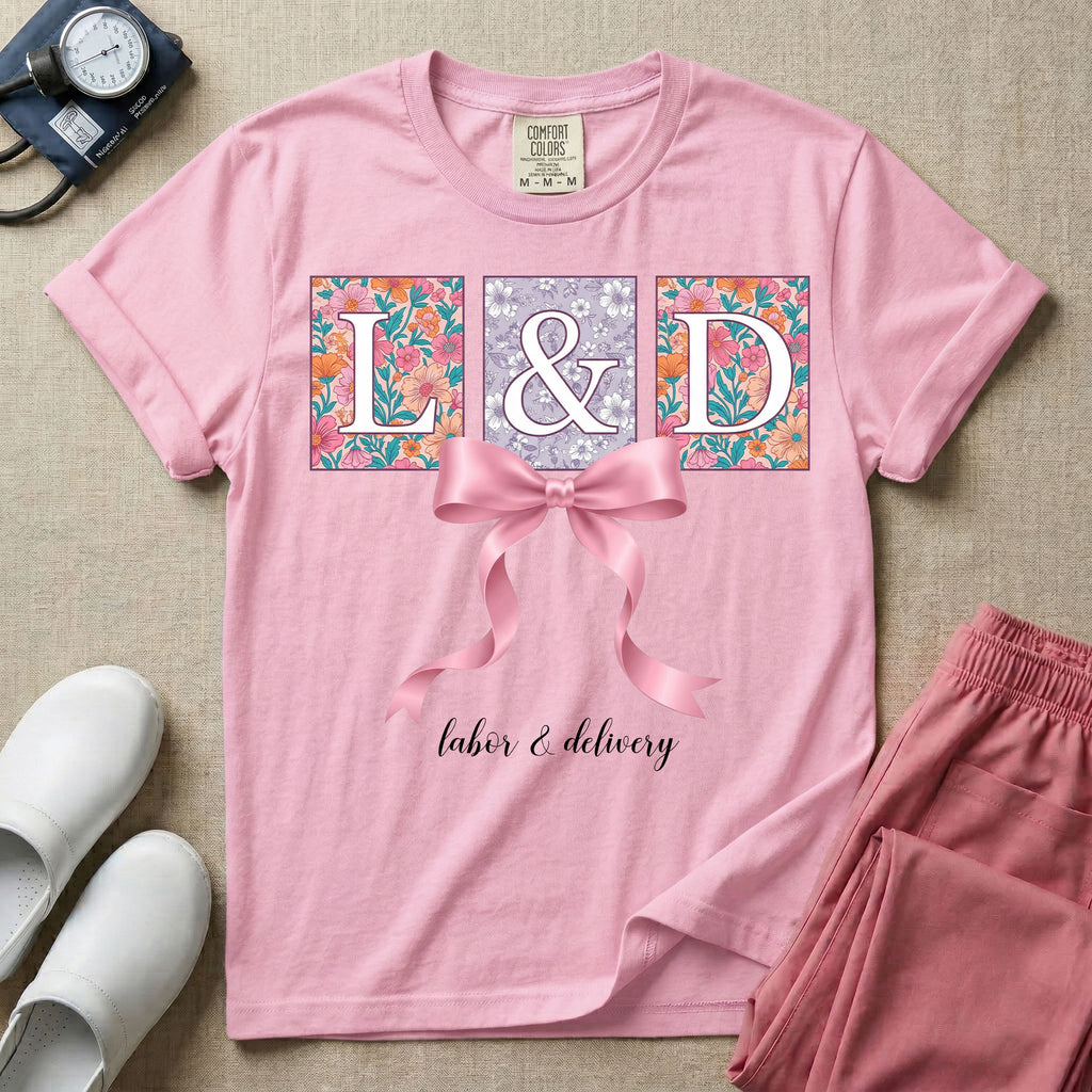 A graphic t-shirt for a Labor and Delivery unit featuring the white serif letters 'L&D' set inside colorful floral square panels, accented with a large graphic pink bow across the middle and the text 'labor & delivery' in delicate white script at the bottom.