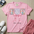 A graphic t-shirt for a Labor and Delivery unit featuring the white serif letters 'L&D' set inside colorful floral square panels, accented with a large graphic pink bow across the middle and the text 'labor & delivery' in delicate white script at the bottom.