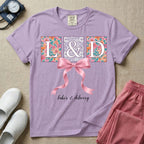 A graphic t-shirt for a Labor and Delivery unit featuring the white serif letters 'L&D' set inside colorful floral square panels, accented with a large graphic pink bow across the middle and the text 'labor & delivery' in delicate white script at the bottom.