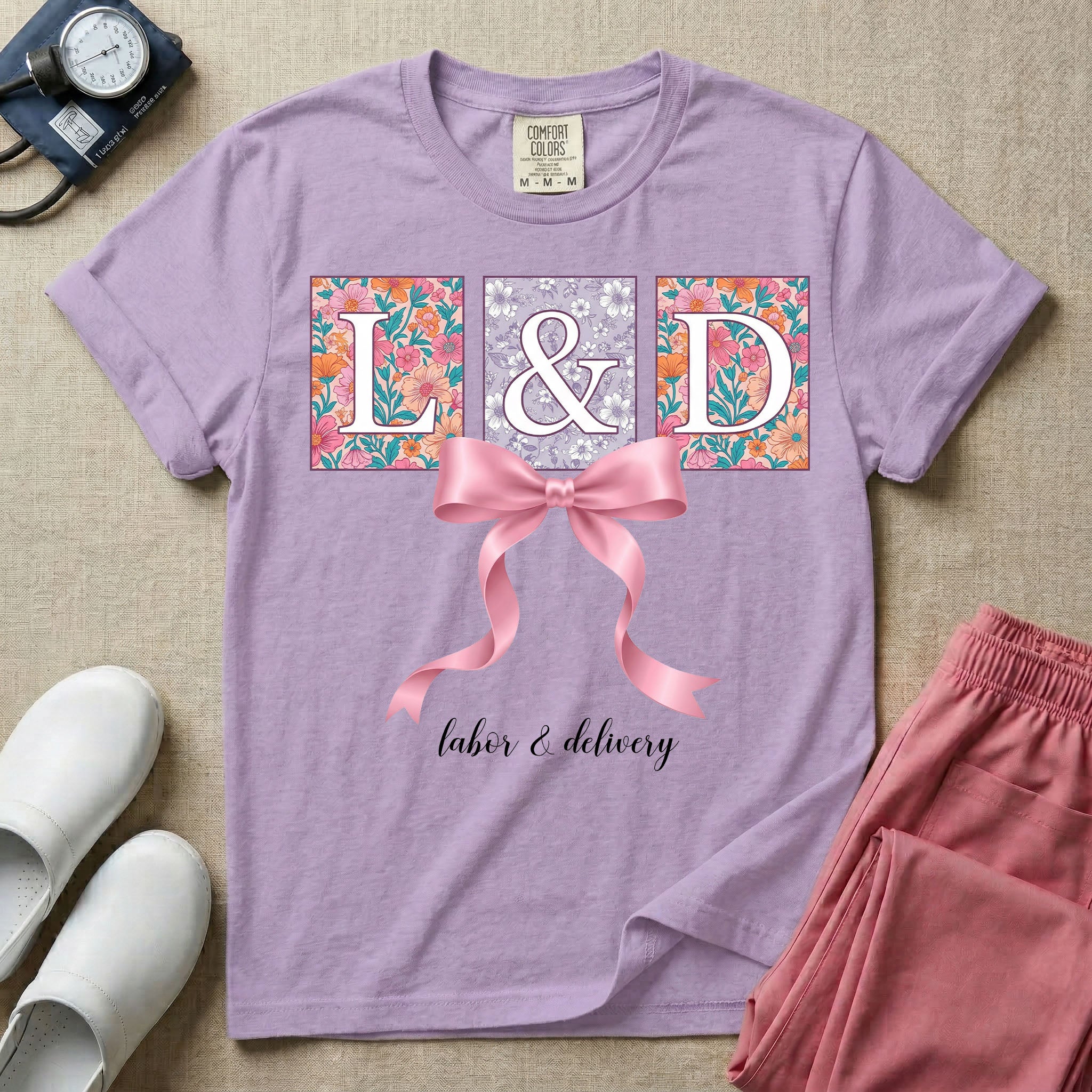 A graphic t-shirt for a Labor and Delivery unit featuring the white serif letters 'L&D' set inside colorful floral square panels, accented with a large graphic pink bow across the middle and the text 'labor & delivery' in delicate white script at the bottom.