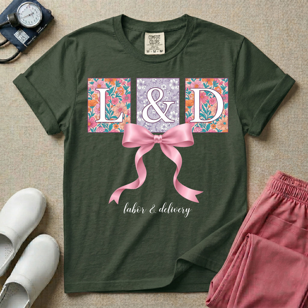 A graphic t-shirt for a Labor and Delivery unit featuring the white serif letters 'L&D' set inside colorful floral square panels, accented with a large graphic pink bow across the middle and the text 'labor & delivery' in delicate white script at the bottom.
