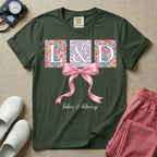 A graphic t-shirt for a Labor and Delivery unit featuring the white serif letters 'L&D' set inside colorful floral square panels, accented with a large graphic pink bow across the middle and the text 'labor & delivery' in delicate white script at the bottom.