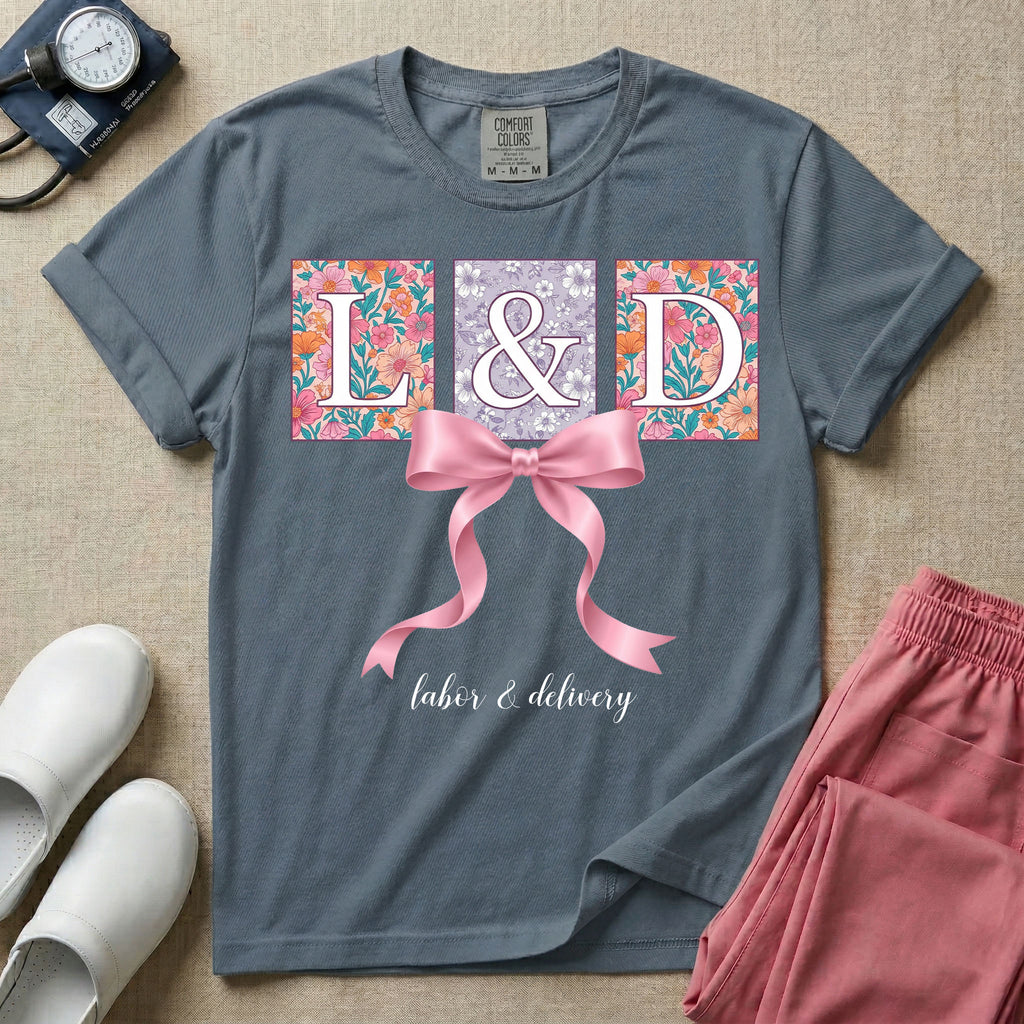 A graphic t-shirt for a Labor and Delivery unit featuring the white serif letters 'L&D' set inside colorful floral square panels, accented with a large graphic pink bow across the middle and the text 'labor & delivery' in delicate white script at the bottom.