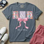 A graphic t-shirt for a Labor and Delivery unit featuring the white serif letters 'L&D' set inside colorful floral square panels, accented with a large graphic pink bow across the middle and the text 'labor & delivery' in delicate white script at the bottom.