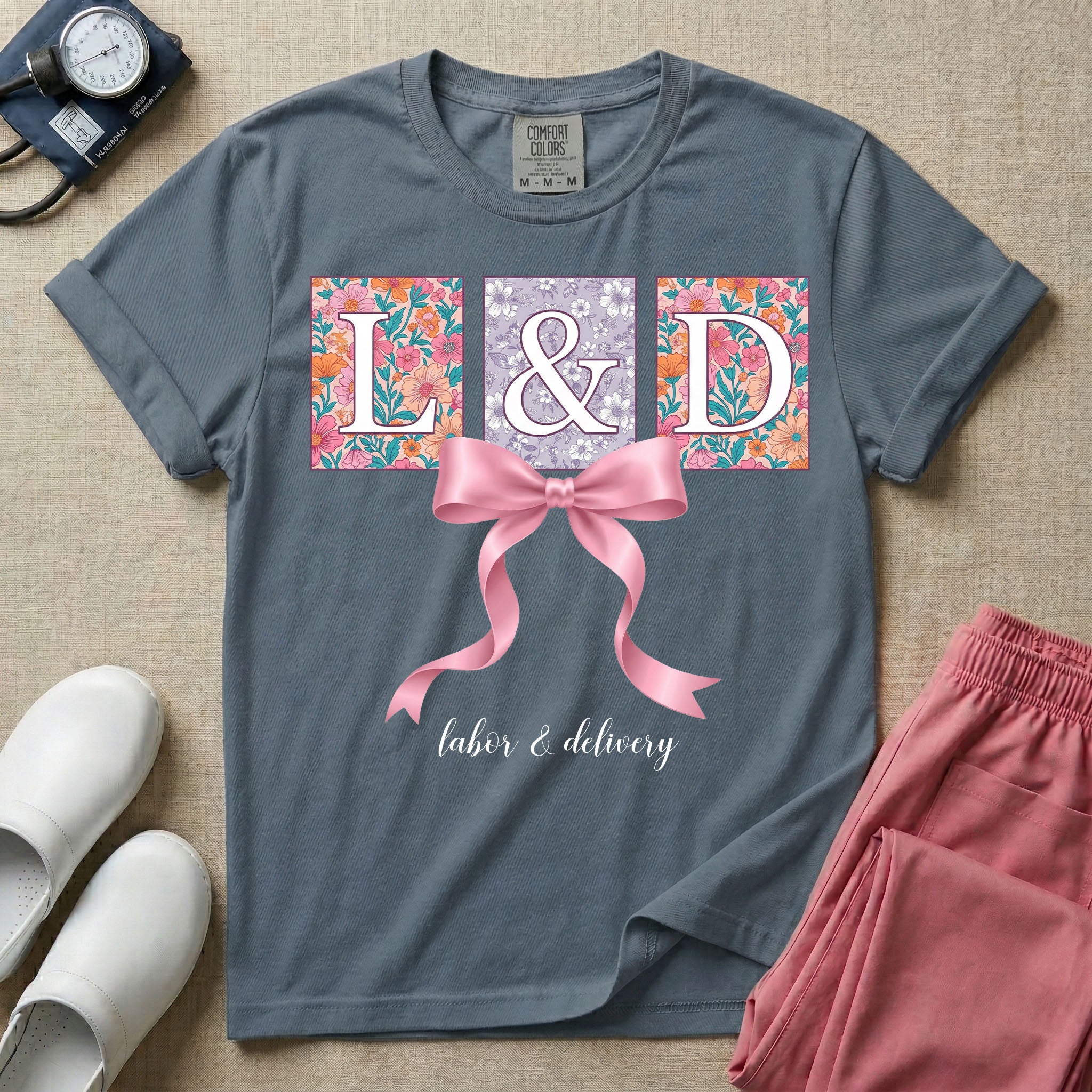 A graphic t-shirt for a Labor and Delivery unit featuring the white serif letters 'L&D' set inside colorful floral square panels, accented with a large graphic pink bow across the middle and the text 'labor & delivery' in delicate white script at the bottom.