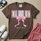 A graphic t-shirt for a Labor and Delivery unit featuring the white serif letters 'L&D' set inside colorful floral square panels, accented with a large graphic pink bow across the middle and the text 'labor & delivery' in delicate white script at the bottom.