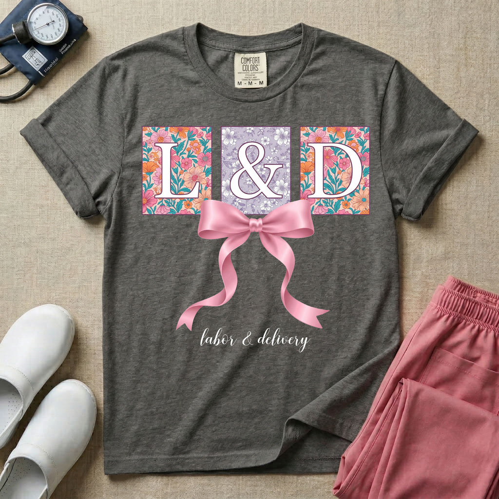 A graphic t-shirt for a Labor and Delivery unit featuring the white serif letters 'L&D' set inside colorful floral square panels, accented with a large graphic pink bow across the middle and the text 'labor & delivery' in delicate white script at the bottom.