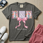 A graphic t-shirt for a Labor and Delivery unit featuring the white serif letters 'L&D' set inside colorful floral square panels, accented with a large graphic pink bow across the middle and the text 'labor & delivery' in delicate white script at the bottom.