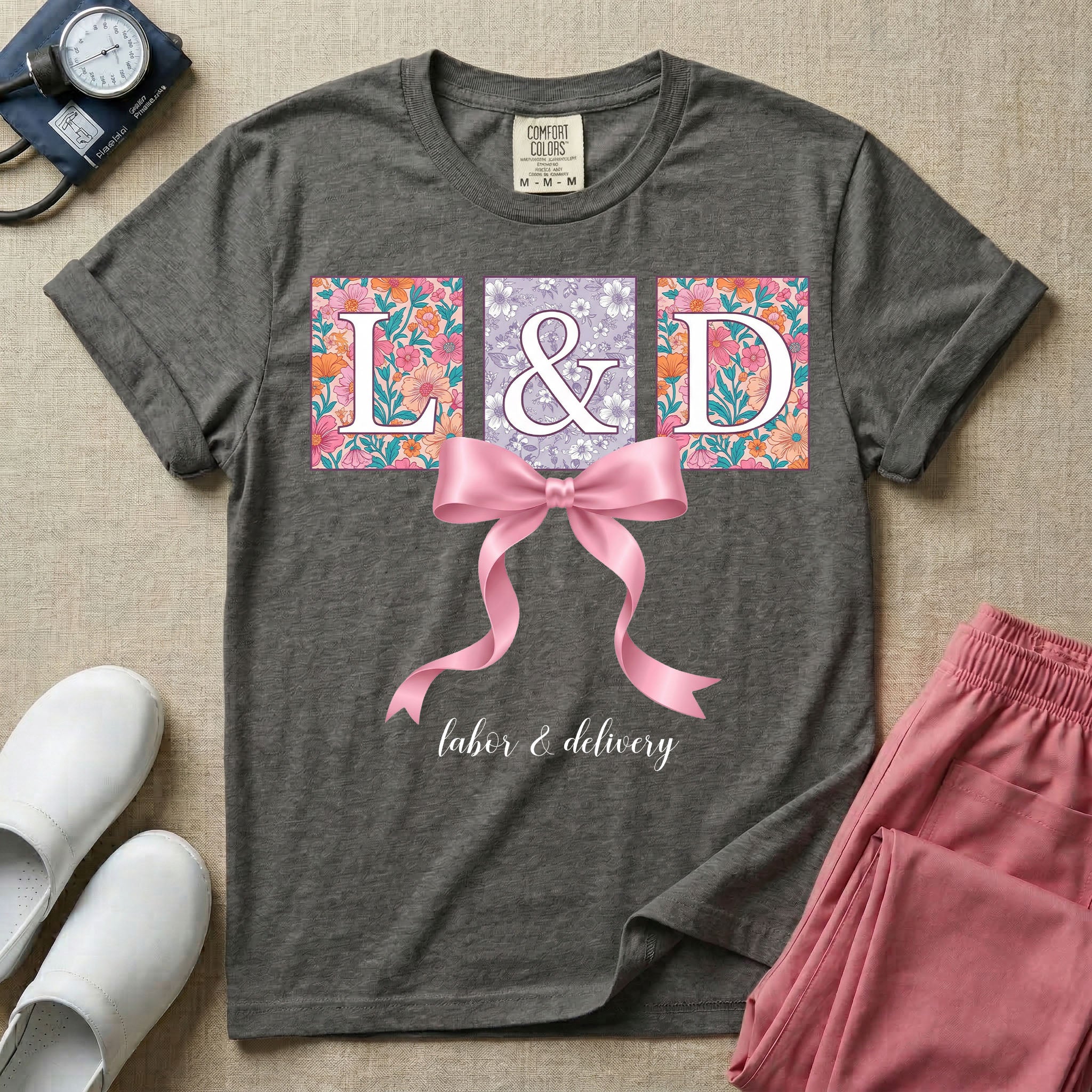 A graphic t-shirt for a Labor and Delivery unit featuring the white serif letters 'L&D' set inside colorful floral square panels, accented with a large graphic pink bow across the middle and the text 'labor & delivery' in delicate white script at the bottom.