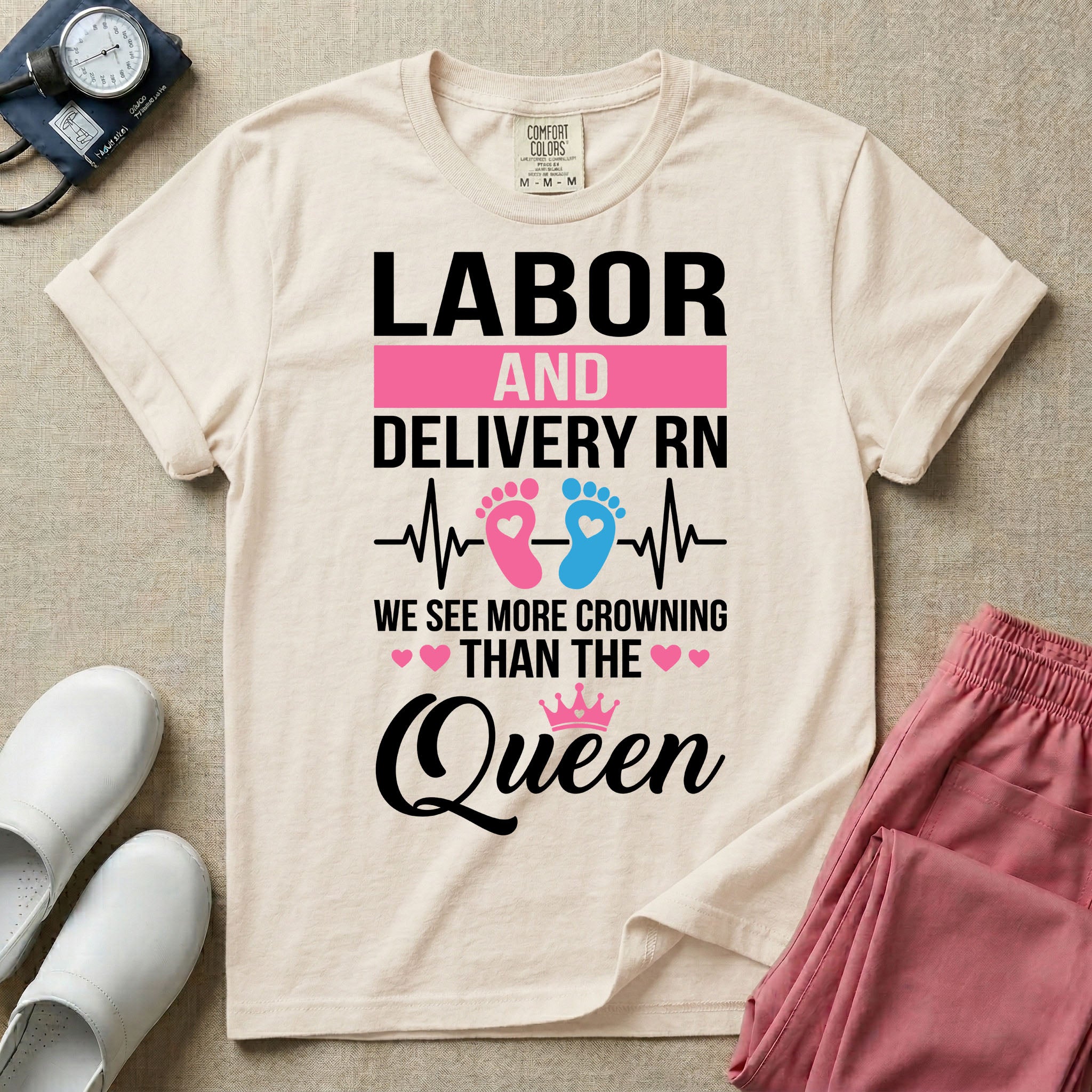 A humorous Labor and Delivery RN design featuring an EKG heartbeat line with pink and blue baby footprints, tiny pink hearts, a pink crown, and the text "LABOR AND DELIVERY RN WE SEE MORE CROWNING THAN THE Queen".