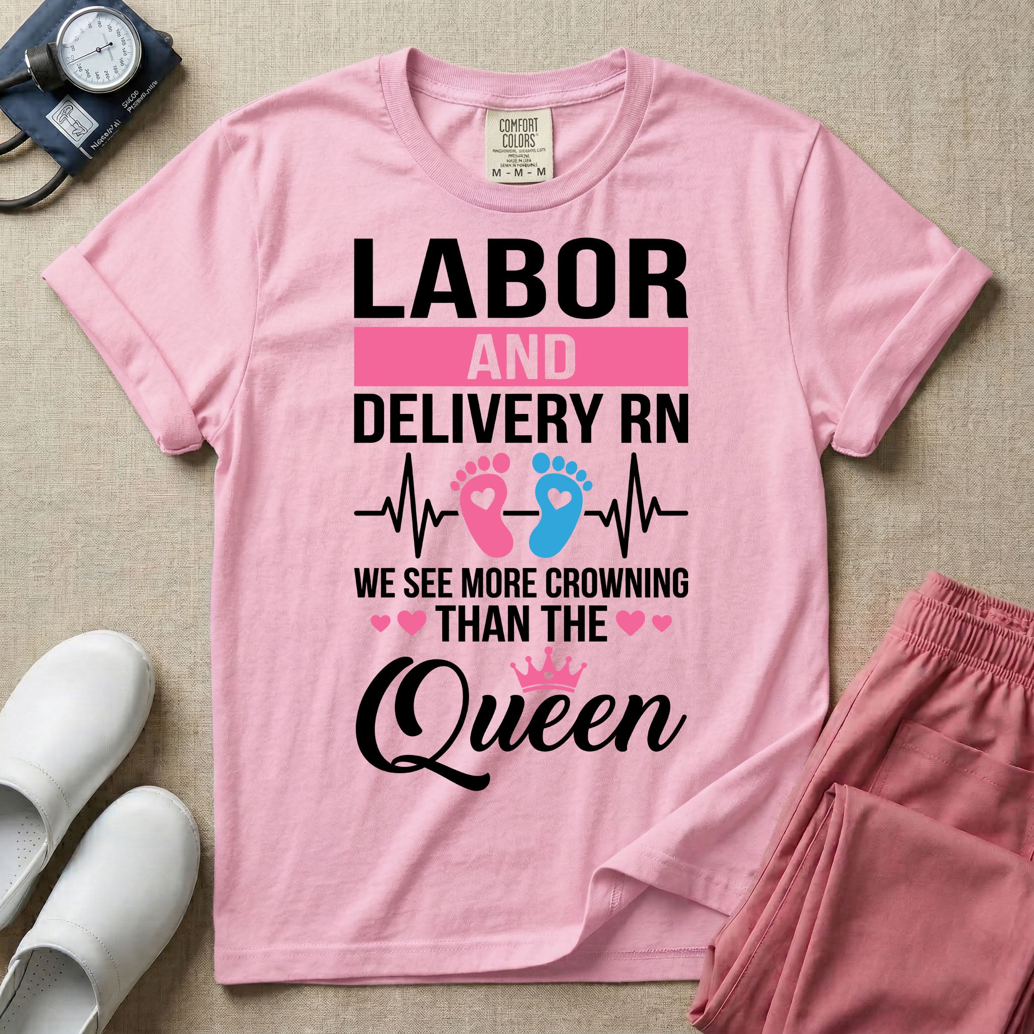 A humorous Labor and Delivery RN design featuring an EKG heartbeat line with pink and blue baby footprints, tiny pink hearts, a pink crown, and the text "LABOR AND DELIVERY RN WE SEE MORE CROWNING THAN THE Queen".