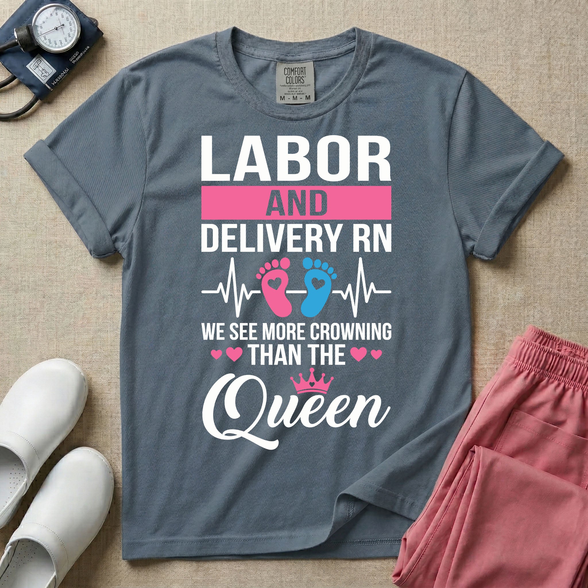 A humorous Labor and Delivery RN design featuring an EKG heartbeat line with pink and blue baby footprints, tiny pink hearts, a pink crown, and the text "LABOR AND DELIVERY RN WE SEE MORE CROWNING THAN THE Queen".