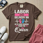 A humorous Labor and Delivery RN design featuring an EKG heartbeat line with pink and blue baby footprints, tiny pink hearts, a pink crown, and the text "LABOR AND DELIVERY RN WE SEE MORE CROWNING THAN THE Queen".