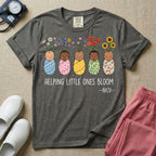A Neonatal Intensive Care Unit nurse design featuring five peacefully sleeping, diverse babies in differently patterned swaddles with vibrant flowers like sunflowers and daisies blooming from the tops of their heads, positioned above the text "HELPING LITTLE ONES BLOOM -NICU-".