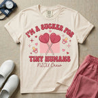 A retro Neonatal Intensive Care Unit nurse design featuring two crossed heart-shaped lollipops over a textured pink square, flanked by two baby pacifiers, sparkling stars, and small hearts, surrounded by the humorous text "I'M A SUCKER FOR TINY HUMANS NICU Crew" in a red and pink groovy font.