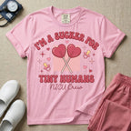 A retro Neonatal Intensive Care Unit nurse design featuring two crossed heart-shaped lollipops over a textured pink square, flanked by two baby pacifiers, sparkling stars, and small hearts, surrounded by the humorous text "I'M A SUCKER FOR TINY HUMANS NICU Crew" in a red and pink groovy font.