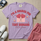 A retro Neonatal Intensive Care Unit nurse design featuring two crossed heart-shaped lollipops over a textured pink square, flanked by two baby pacifiers, sparkling stars, and small hearts, surrounded by the humorous text "I'M A SUCKER FOR TINY HUMANS NICU Crew" in a red and pink groovy font.