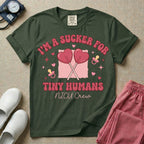 A retro Neonatal Intensive Care Unit nurse design featuring two crossed heart-shaped lollipops over a textured pink square, flanked by two baby pacifiers, sparkling stars, and small hearts, surrounded by the humorous text "I'M A SUCKER FOR TINY HUMANS NICU Crew" in a red and pink groovy font.