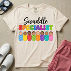 A graphic featuring the word Swaddle in white cursive above the word SPECIALIST in bright, colorful bubble letters, resting over a row of eight diverse babies wrapped in colorful patterned swaddles, designed for NICU, labor and delivery, postpartum, and pediatric nurses.