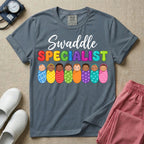 A graphic featuring the word Swaddle in white cursive above the word SPECIALIST in bright, colorful bubble letters, resting over a row of eight diverse babies wrapped in colorful patterned swaddles, designed for NICU, labor and delivery, postpartum, and pediatric nurses.