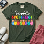 A graphic featuring the word Swaddle in white cursive above the word SPECIALIST in bright, colorful bubble letters, resting over a row of eight diverse babies wrapped in colorful patterned swaddles, designed for NICU, labor and delivery, postpartum, and pediatric nurses.