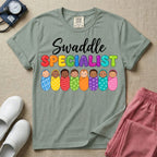 A graphic featuring the word Swaddle in white cursive above the word SPECIALIST in bright, colorful bubble letters, resting over a row of eight diverse babies wrapped in colorful patterned swaddles, designed for NICU, labor and delivery, postpartum, and pediatric nurses.