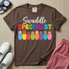 A graphic featuring the word Swaddle in white cursive above the word SPECIALIST in bright, colorful bubble letters, resting over a row of eight diverse babies wrapped in colorful patterned swaddles, designed for NICU, labor and delivery, postpartum, and pediatric nurses.