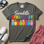A graphic featuring the word Swaddle in white cursive above the word SPECIALIST in bright, colorful bubble letters, resting over a row of eight diverse babies wrapped in colorful patterned swaddles, designed for NICU, labor and delivery, postpartum, and pediatric nurses.