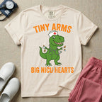 A funny neonatal intensive care unit t-shirt featuring an illustration of a green T-Rex dinosaur wearing a nurse cap and stethoscope, juggling a baby bottle and pacifiers, surrounded by distressed orange text that reads "TINY ARMS" at the top and "BIG NICU HEARTS" at the bottom.