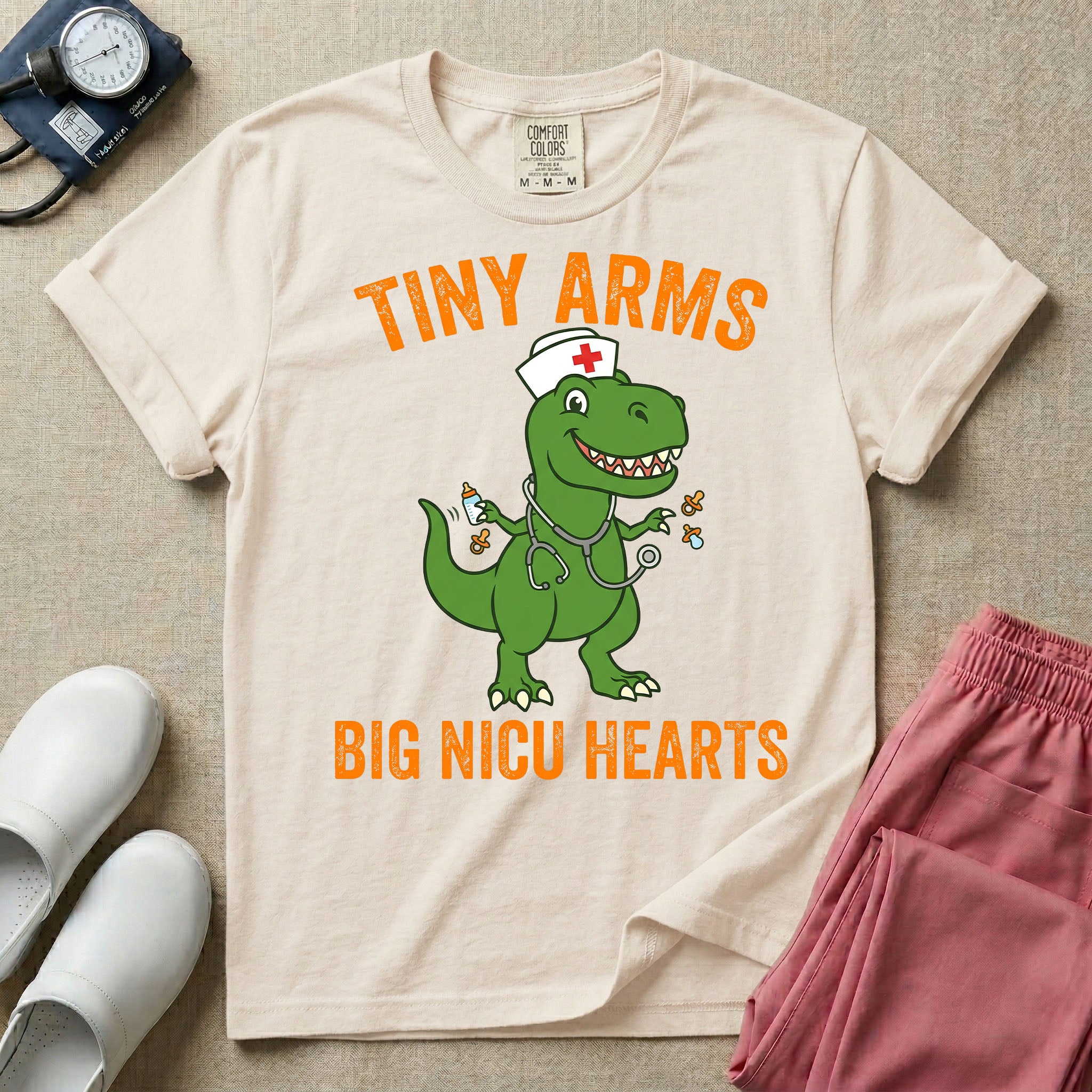 A funny neonatal intensive care unit t-shirt featuring an illustration of a green T-Rex dinosaur wearing a nurse cap and stethoscope, juggling a baby bottle and pacifiers, surrounded by distressed orange text that reads "TINY ARMS" at the top and "BIG NICU HEARTS" at the bottom.