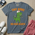 A funny neonatal intensive care unit t-shirt featuring an illustration of a green T-Rex dinosaur wearing a nurse cap and stethoscope, juggling a baby bottle and pacifiers, surrounded by distressed orange text that reads "TINY ARMS" at the top and "BIG NICU HEARTS" at the bottom.
