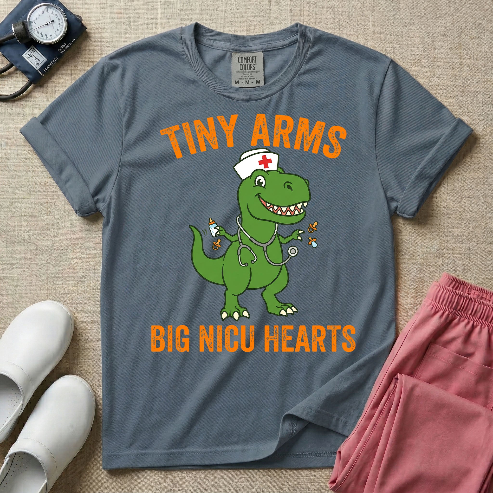 A funny neonatal intensive care unit t-shirt featuring an illustration of a green T-Rex dinosaur wearing a nurse cap and stethoscope, juggling a baby bottle and pacifiers, surrounded by distressed orange text that reads "TINY ARMS" at the top and "BIG NICU HEARTS" at the bottom.