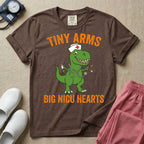 A funny neonatal intensive care unit t-shirt featuring an illustration of a green T-Rex dinosaur wearing a nurse cap and stethoscope, juggling a baby bottle and pacifiers, surrounded by distressed orange text that reads "TINY ARMS" at the top and "BIG NICU HEARTS" at the bottom.