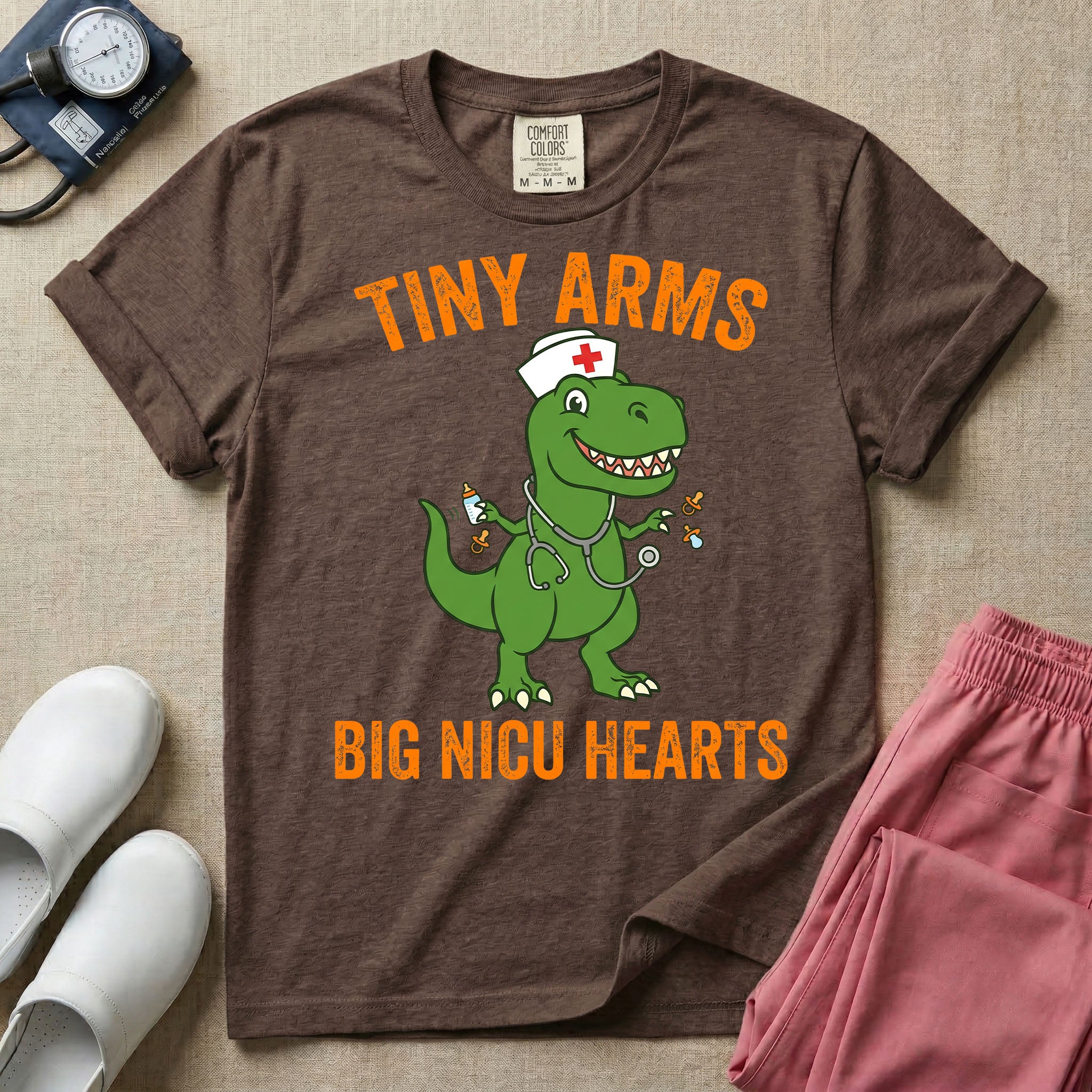 A funny neonatal intensive care unit t-shirt featuring an illustration of a green T-Rex dinosaur wearing a nurse cap and stethoscope, juggling a baby bottle and pacifiers, surrounded by distressed orange text that reads "TINY ARMS" at the top and "BIG NICU HEARTS" at the bottom.