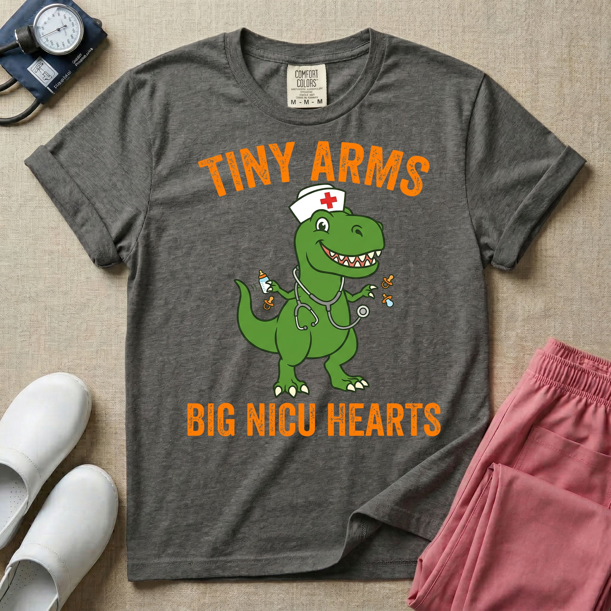 A funny neonatal intensive care unit t-shirt featuring an illustration of a green T-Rex dinosaur wearing a nurse cap and stethoscope, juggling a baby bottle and pacifiers, surrounded by distressed orange text that reads "TINY ARMS" at the top and "BIG NICU HEARTS" at the bottom.