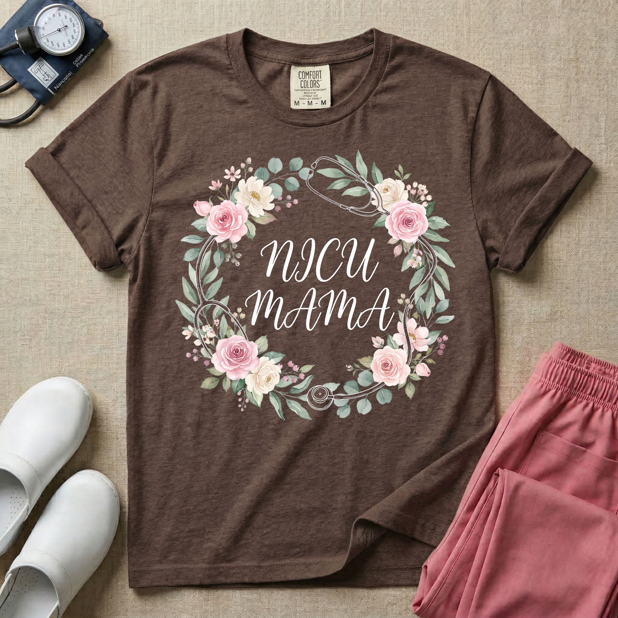 A t-shirt featuring a boho floral wreath composed of pink and cream roses intertwined with a stethoscope, surrounding the text NICU MAMA in elegant typography.