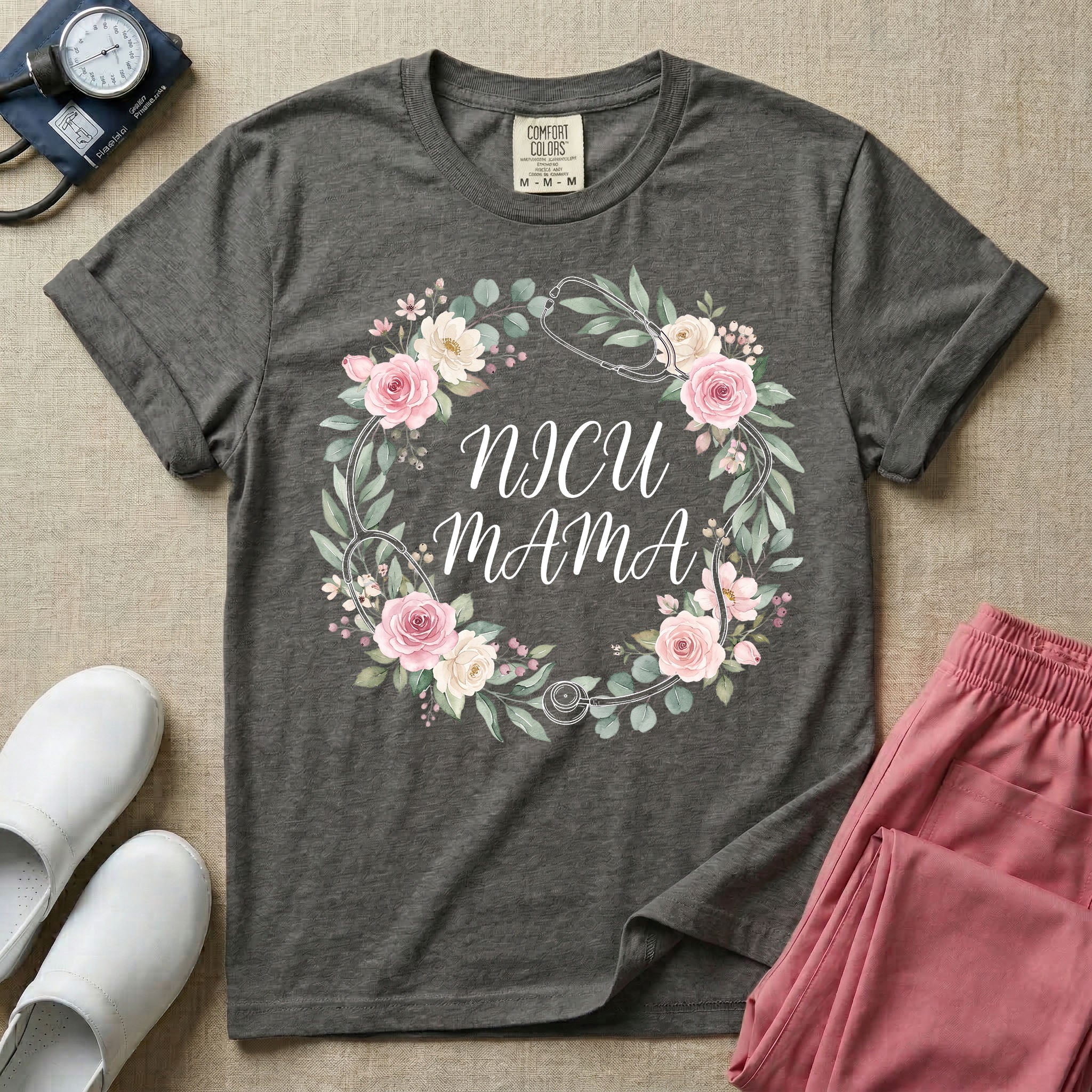 A t-shirt featuring a boho floral wreath composed of pink and cream roses intertwined with a stethoscope, surrounding the text NICU MAMA in elegant typography.