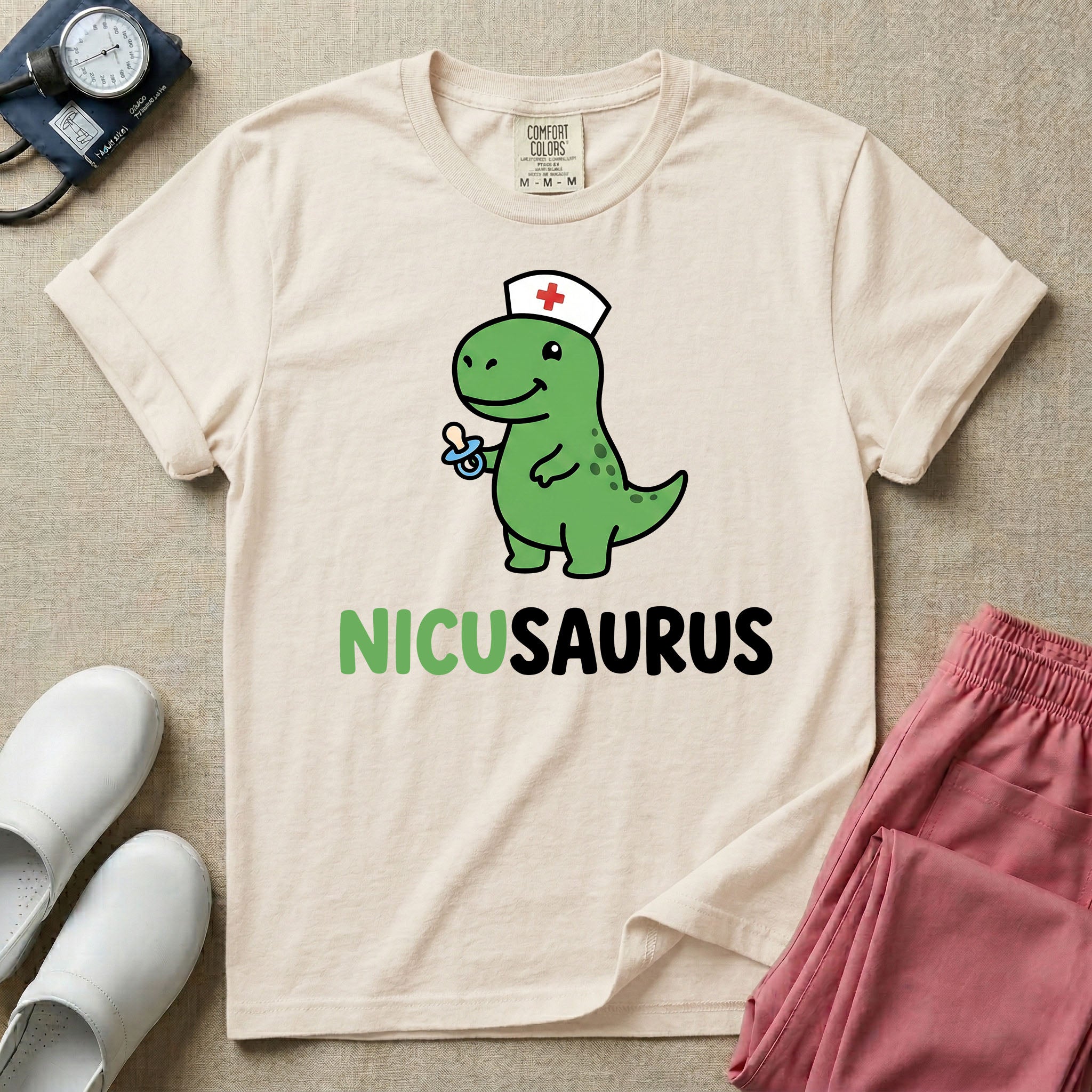 A cute t-shirt for NICU nurses featuring a cartoon green dinosaur wearing a nurse hat with a red cross and holding a blue pacifier above the funny text NICUSAURUS.