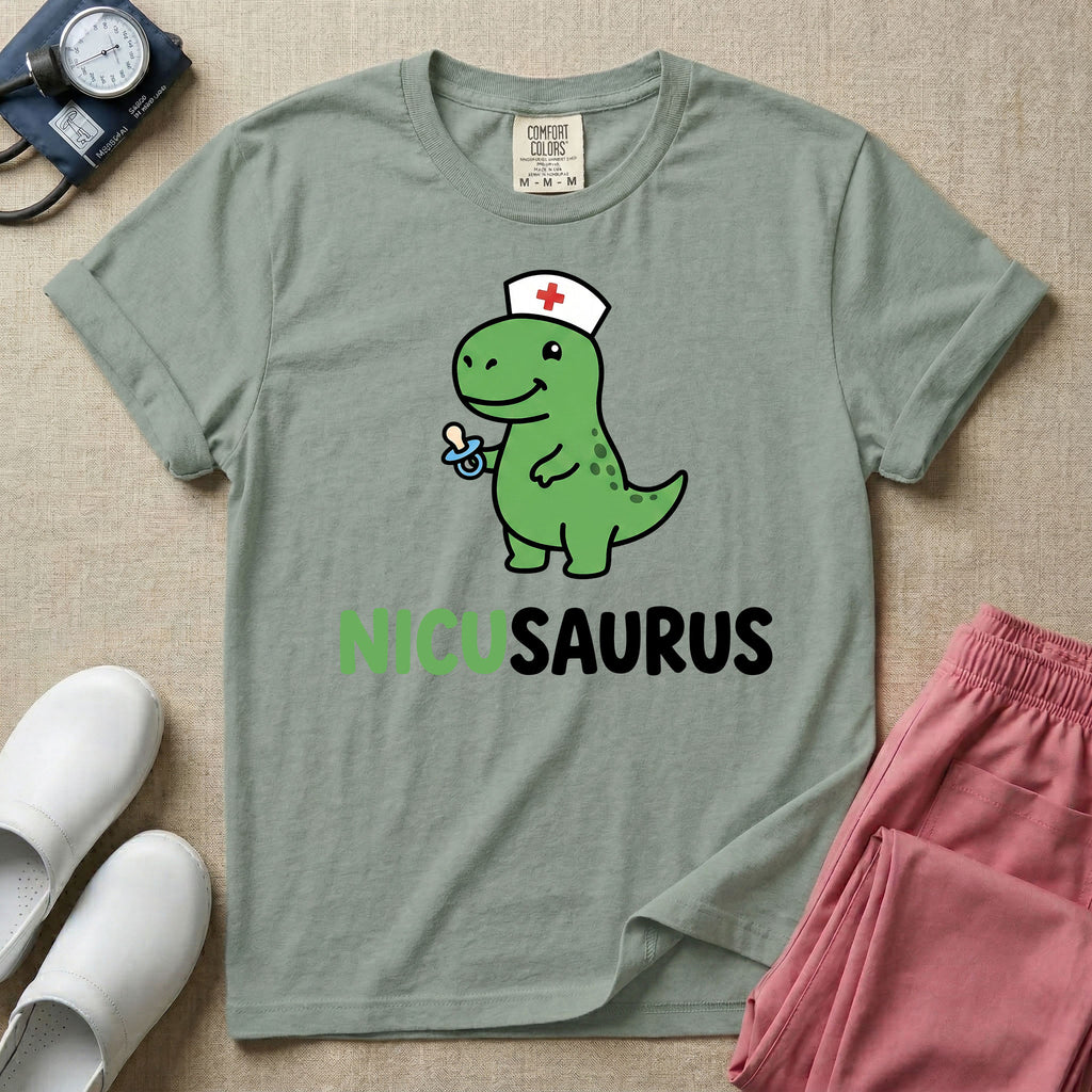 A cute t-shirt for NICU nurses featuring a cartoon green dinosaur wearing a nurse hat with a red cross and holding a blue pacifier above the funny text NICUSAURUS.