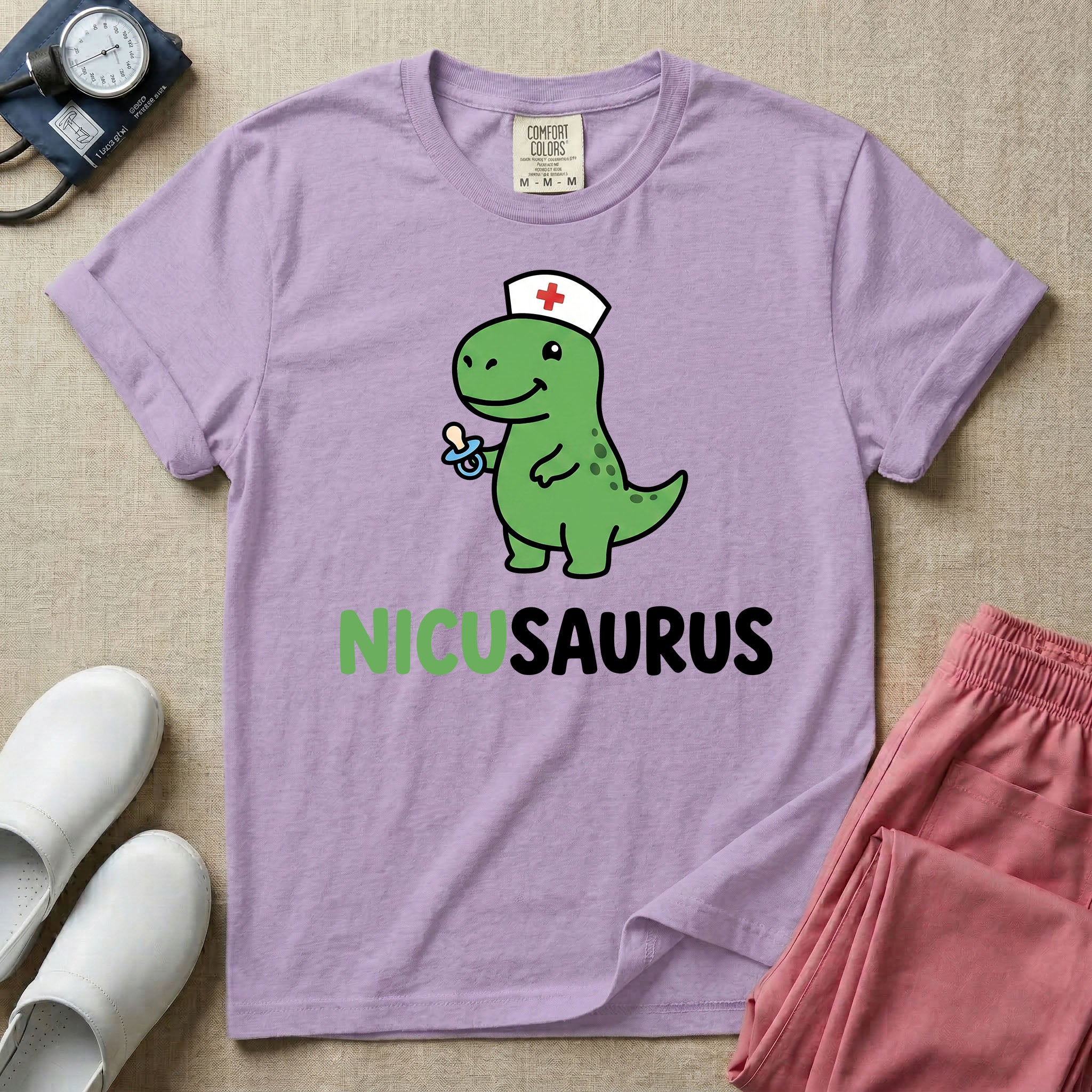 A cute t-shirt for NICU nurses featuring a cartoon green dinosaur wearing a nurse hat with a red cross and holding a blue pacifier above the funny text NICUSAURUS.