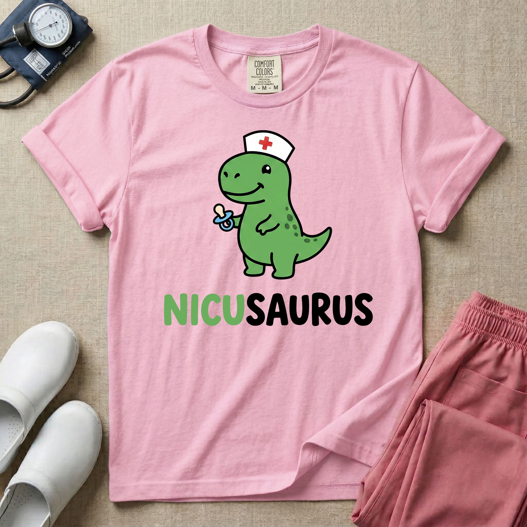A cute t-shirt for NICU nurses featuring a cartoon green dinosaur wearing a nurse hat with a red cross and holding a blue pacifier above the funny text NICUSAURUS.